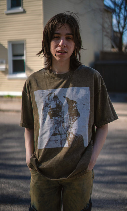 Person wearing a green t-shirt with a graphic design, standing on a street.