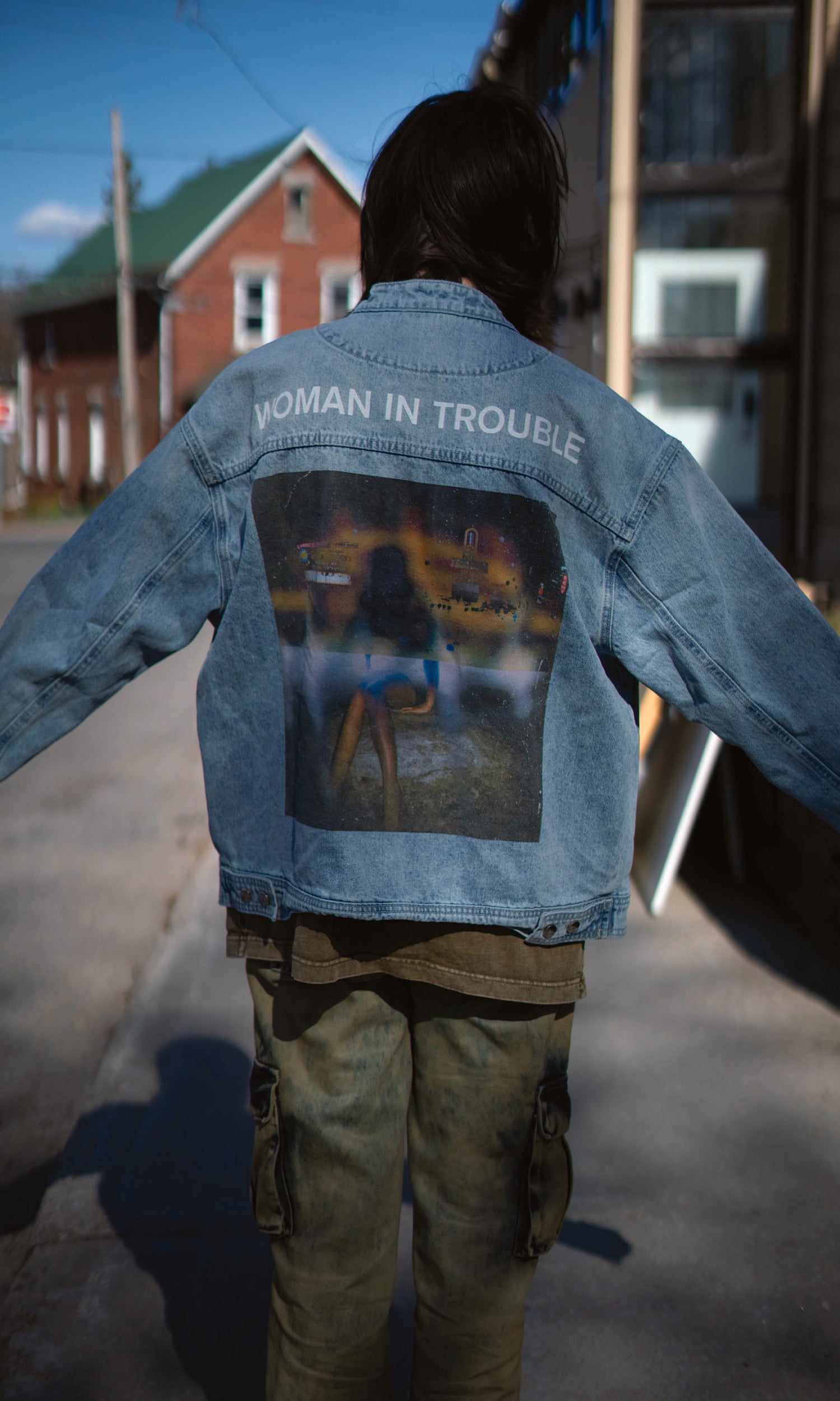 Person wearing a denim jacket with a graphic design on the back, standing outdoors.